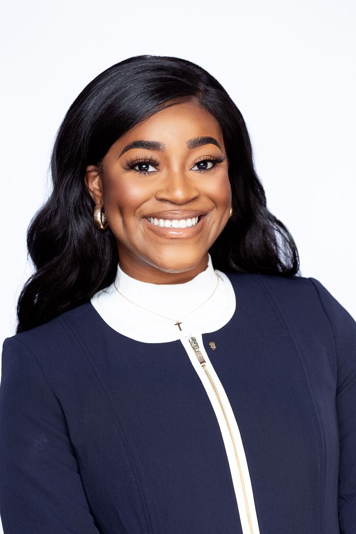 Smiling professional woman in navy blazer with white turtleneck, long black hair, hoop earrings, on white background.