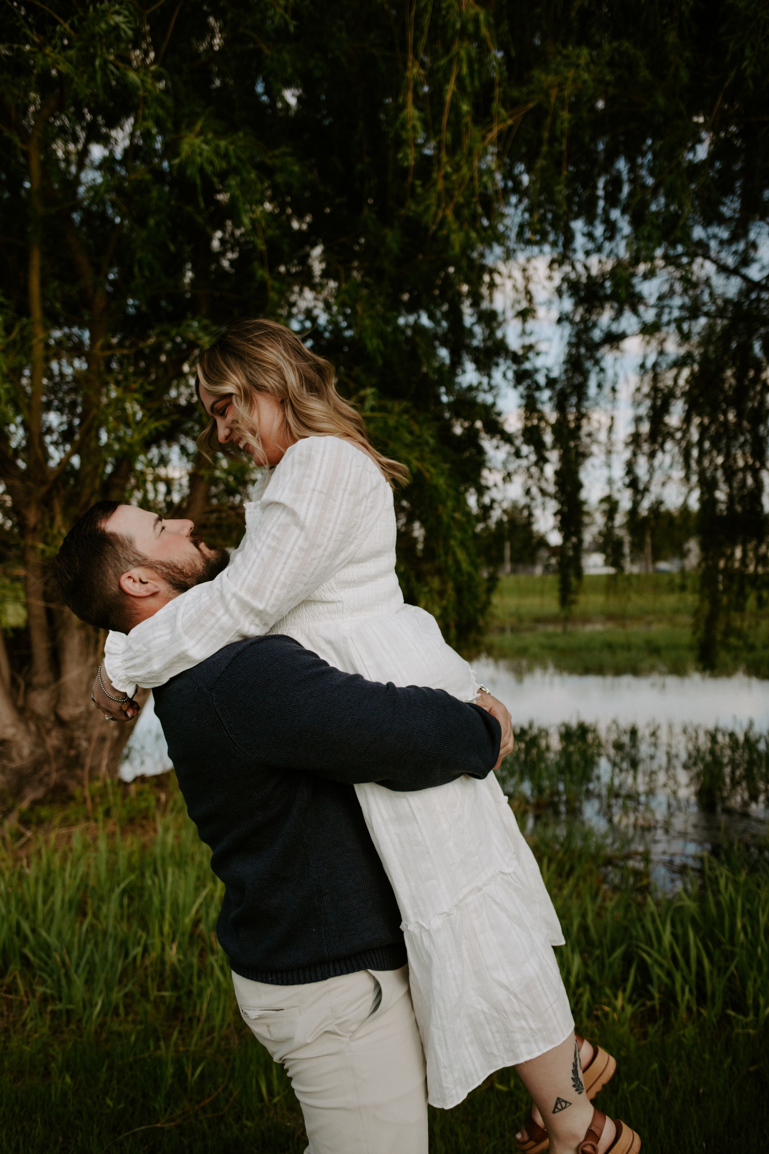 Man lifting woman in white dress outdoors near pond with lush green trees in romantic engagement photo.