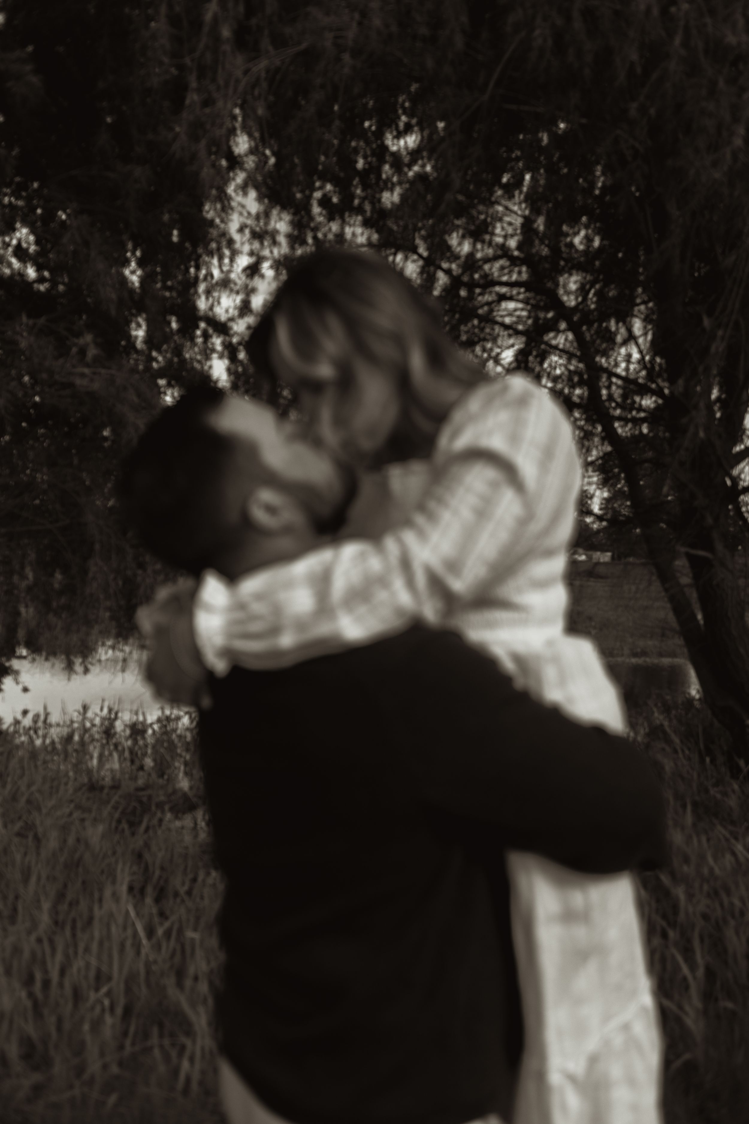 Black and white photo of a couple embracing and kissing near trees by a lake.