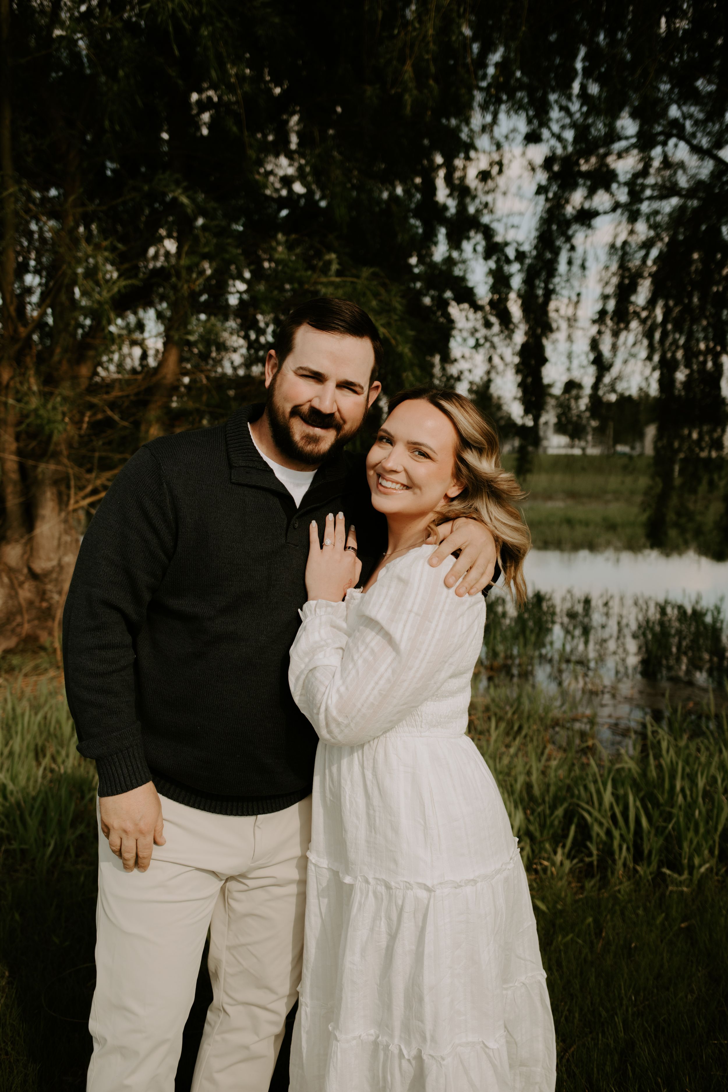 Smiling couple embraces outdoors near a pond, woman in white dress showing engagement ring, man in black sweater.