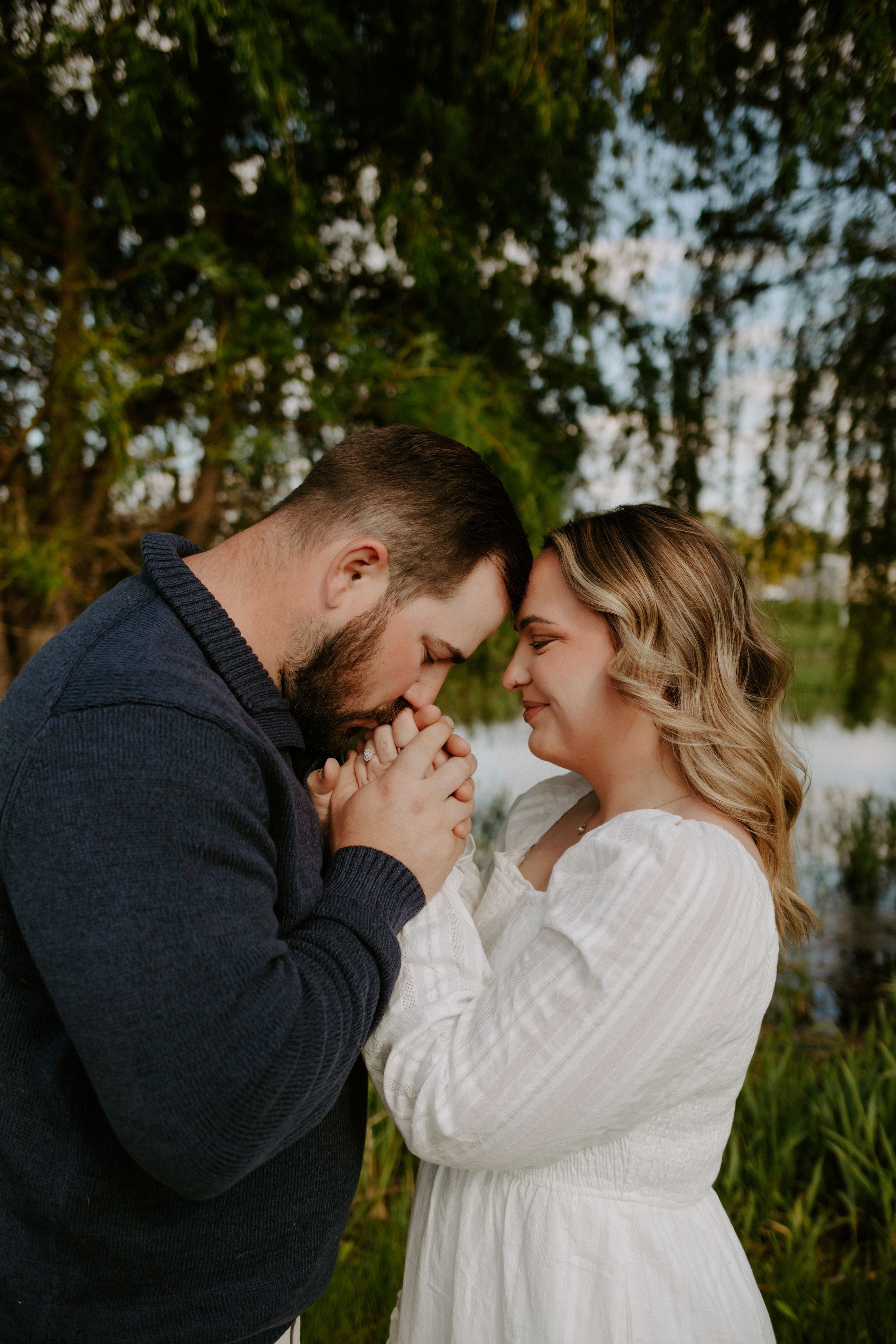 Couple holding hands and touching foreheads tenderly near a pond in a romantic outdoor engagement session.