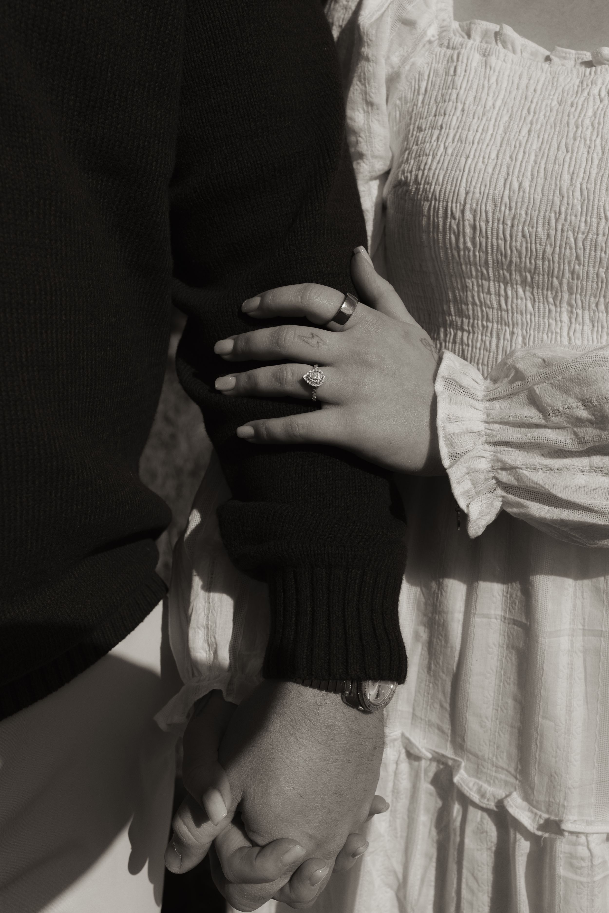 Black and white photo of an engaged couple holding hands, showcasing a diamond engagement ring on the woman's finger.