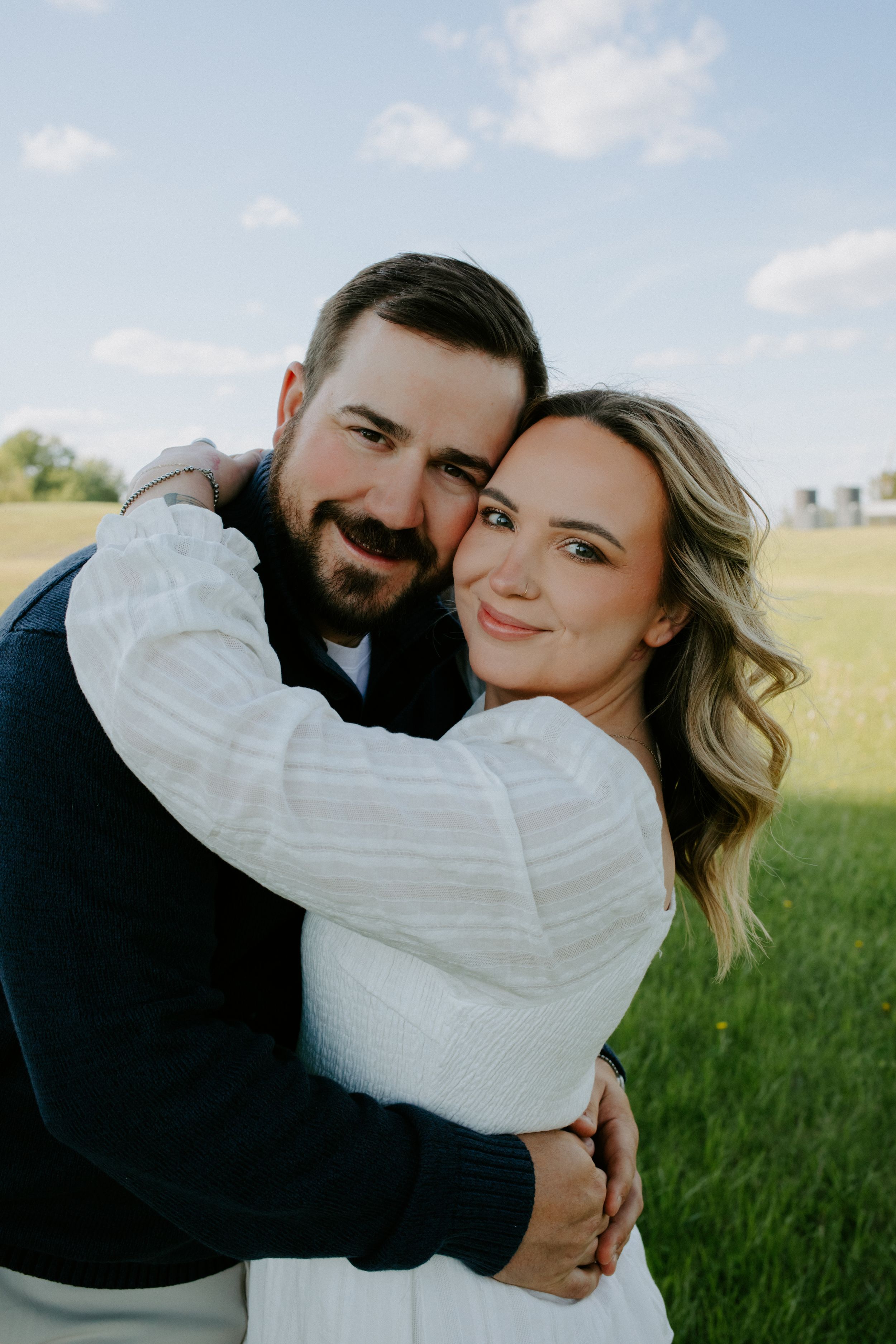 A happy couple embraces outdoors in a green field, smiling warmly in a romantic engagement photo session.