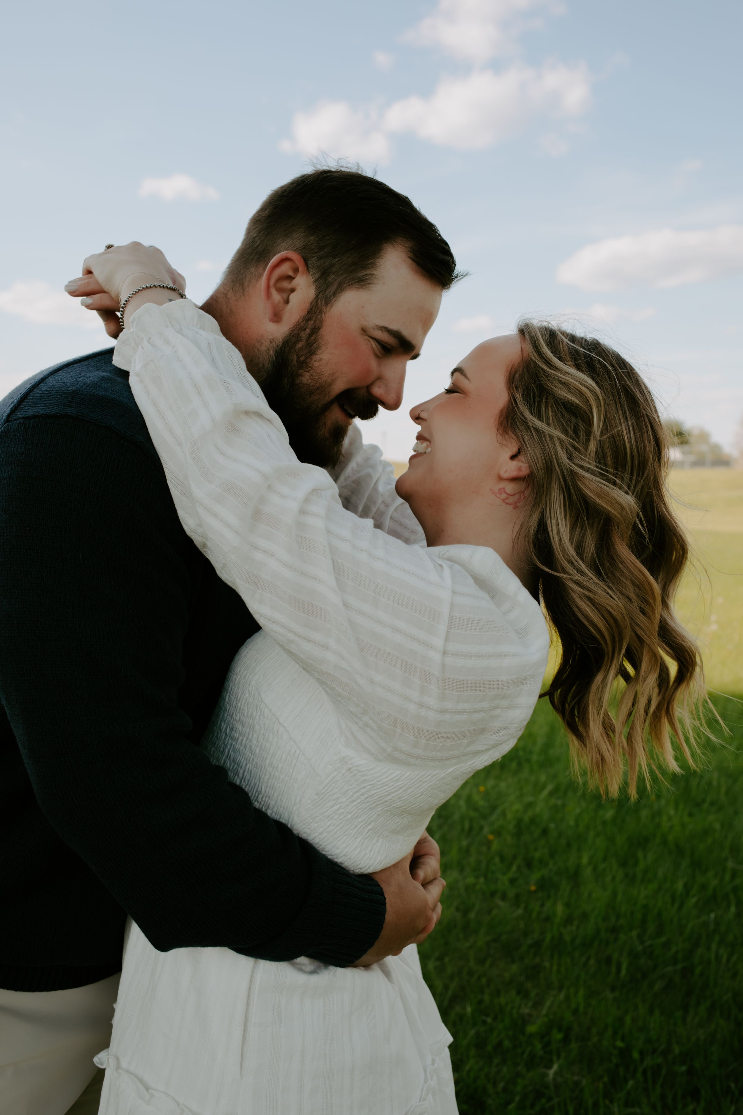 Couple embracing and smiling at each other in a green field under a blue sky.