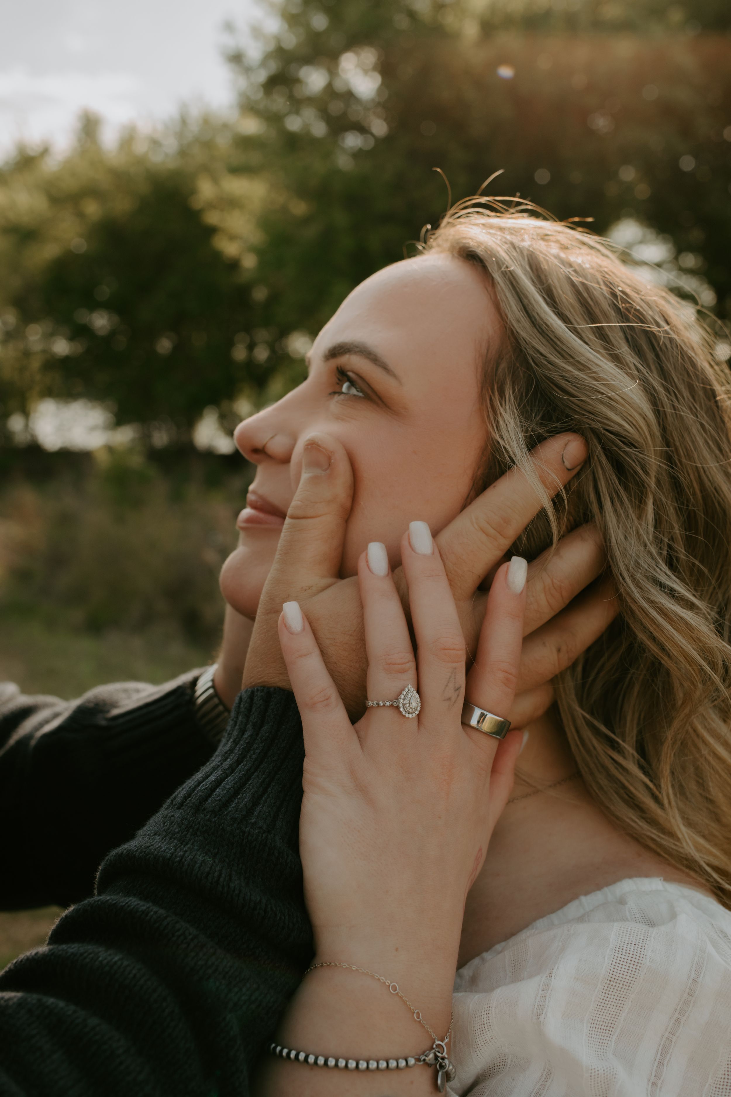 A smiling blonde woman gazes upward as her partner's hands gently cup her face, highlighting her engagement ring outdoors.