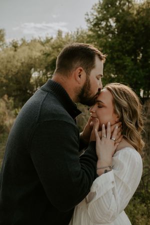 Man tenderly kisses womans forehead while cradling her face, both in elegant outdoor engagement photo.