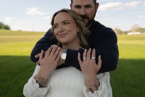 Couple embracing outdoors, woman smiling with engagement ring, man hugging her from behind on green field.