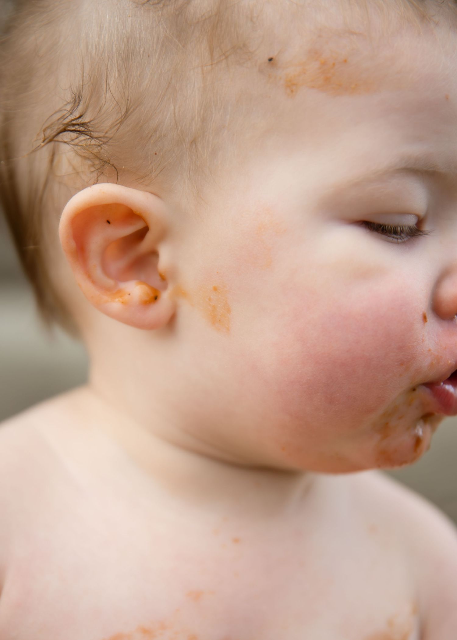 Messy baby with orange food smeared on face, cheeks, and ear after mealtime.