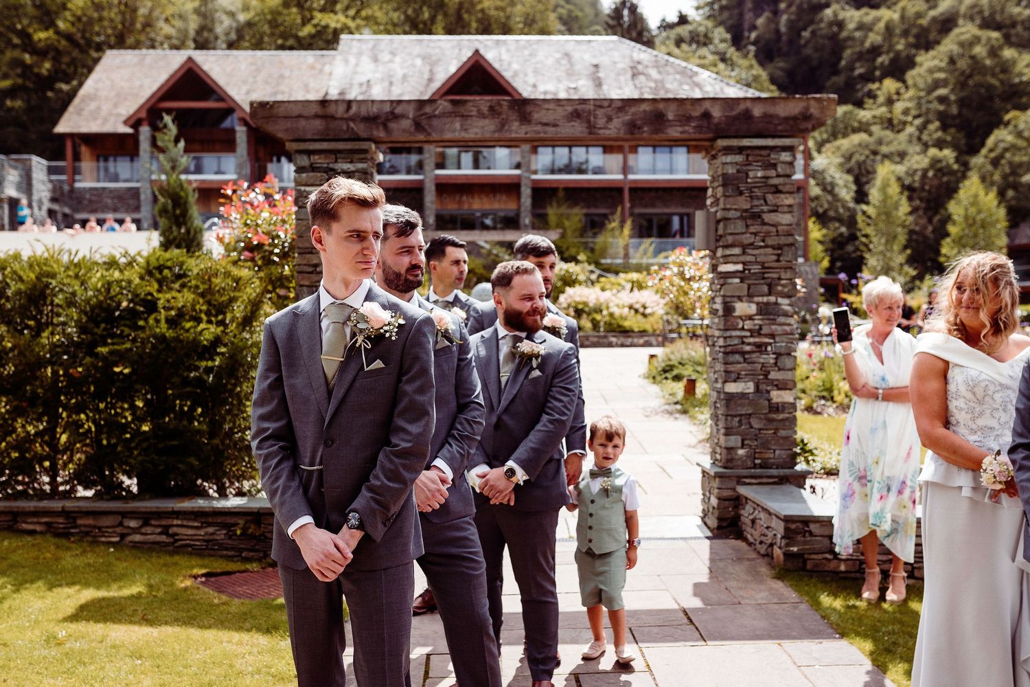 Groomsmen in dark suits stand outdoors at a wedding venue surrounded by lush greenery on a sunny day.