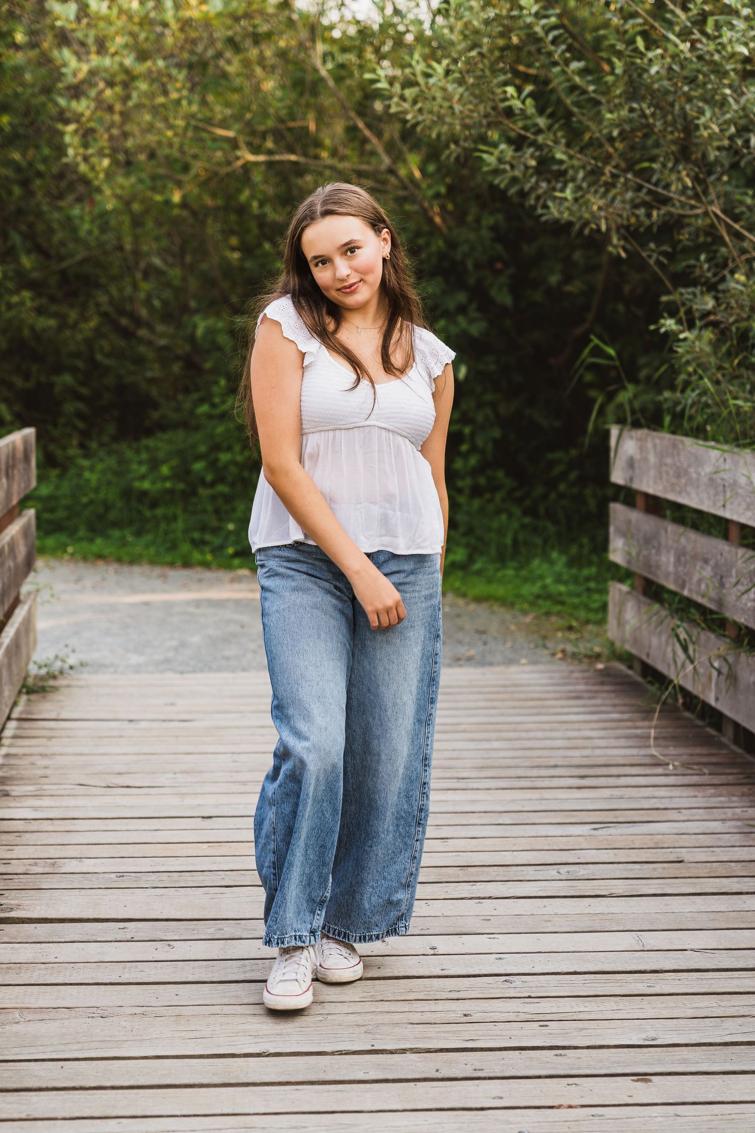 Young woman in white top and wide-leg jeans standing on a wooden bridge surrounded by lush greenery.