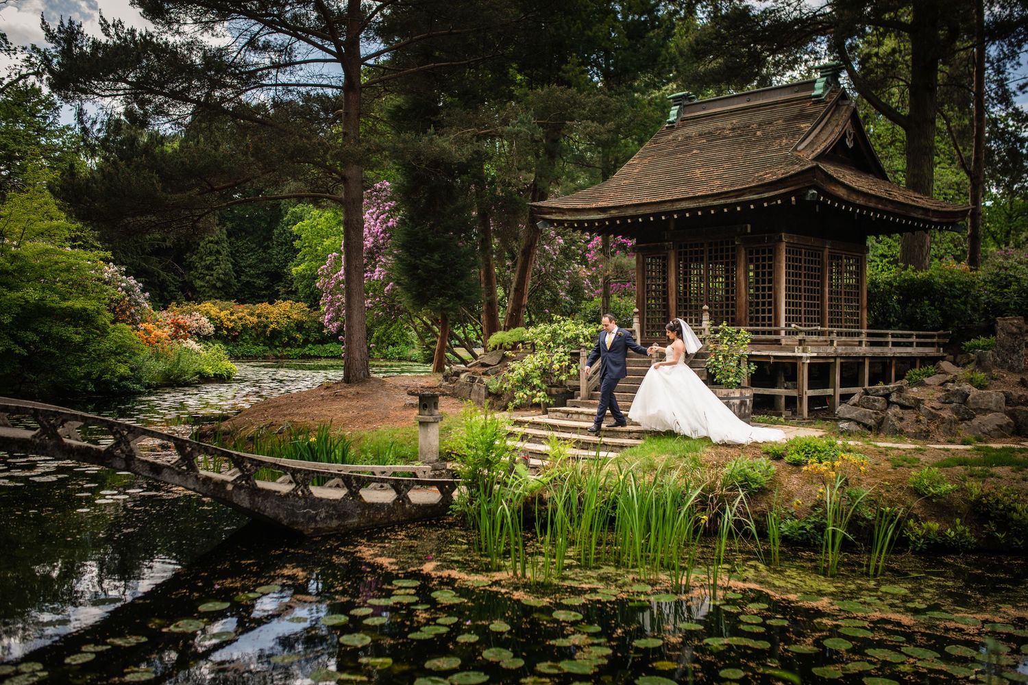 Bride and groom pose near a Japanese pagoda by a serene pond in a lush garden wedding setting.