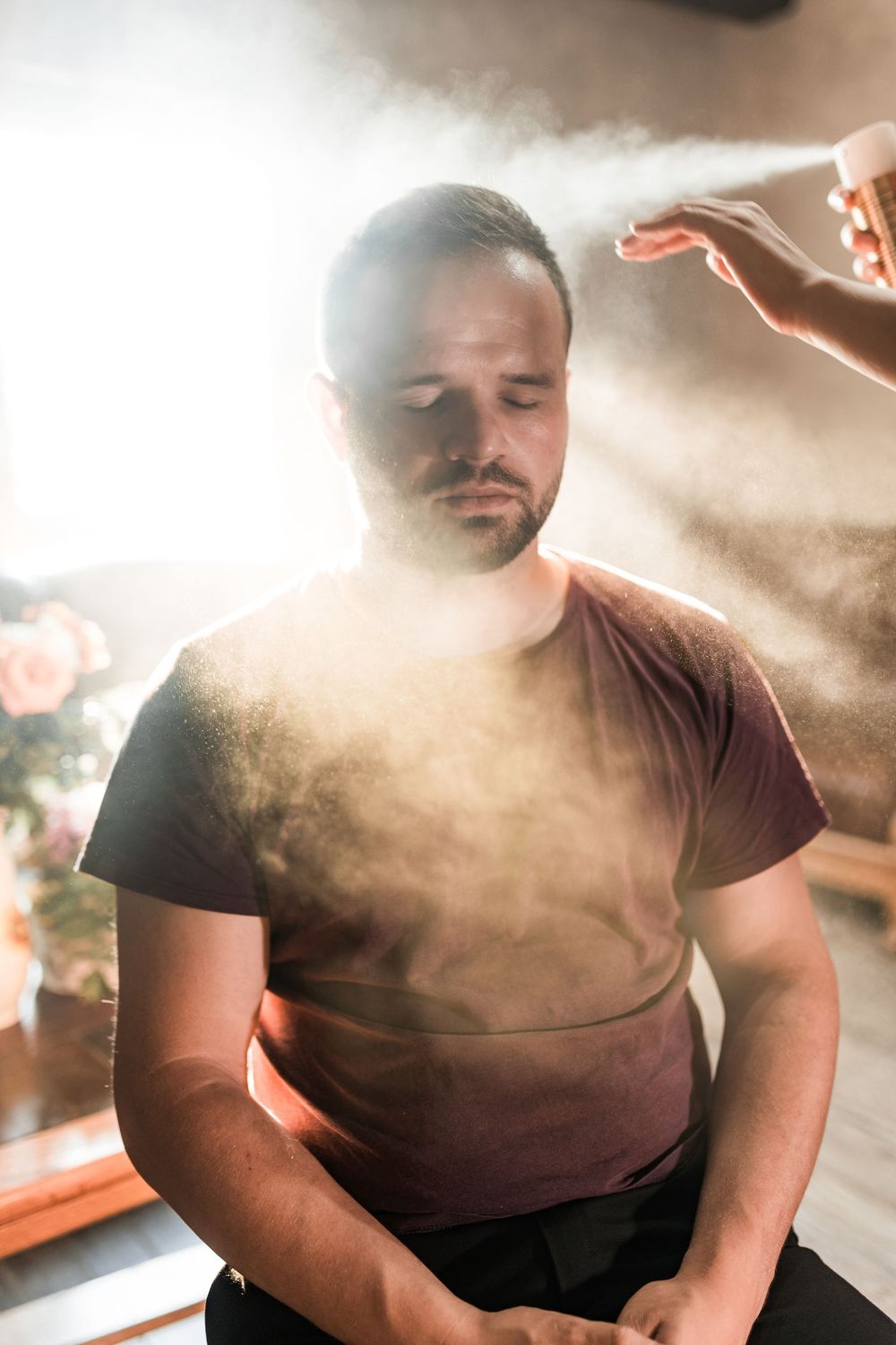 Man meditating with mist spray in sunlit room, eyes closed, wearing burgundy t-shirt.