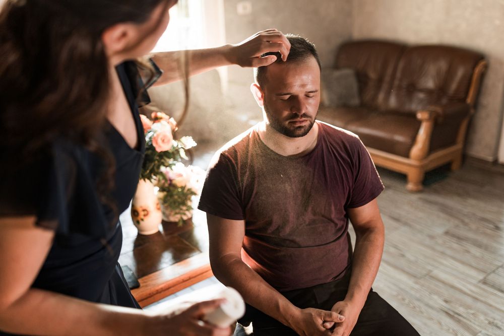 Woman placing hand on seated mans head during indoor healing session.