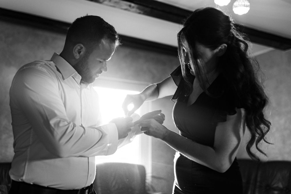 Black and white photo of groom placing ring on brides finger in romantic backlit setting.