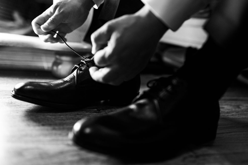 A groom tying his shiny black dress shoes in a black and white wedding preparation photo.