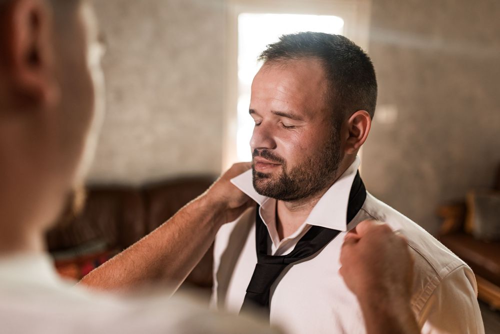 Groom getting his black tie adjusted by a groomsman while preparing for his wedding ceremony.