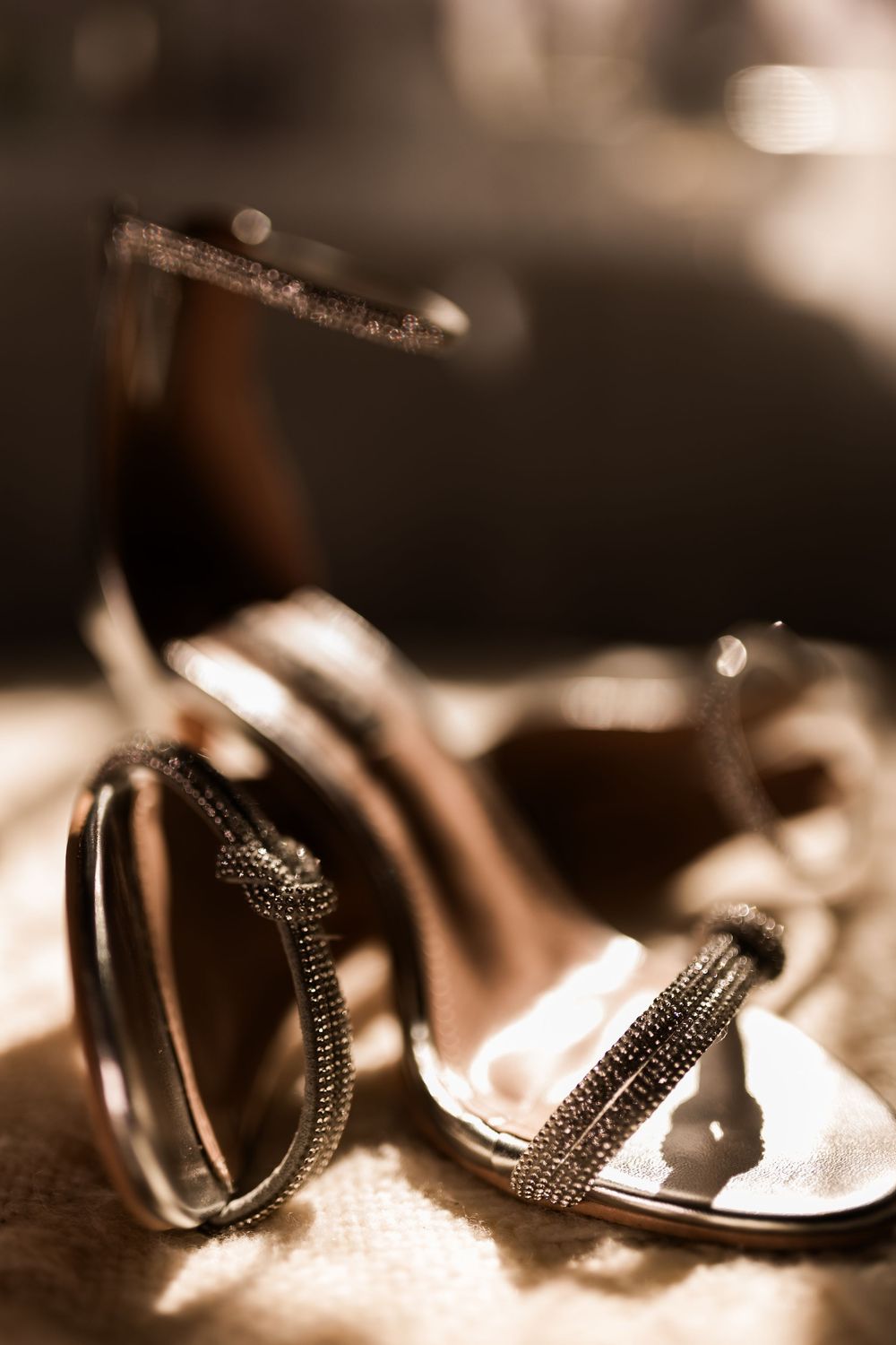 Close-up of elegant silver bridal heels with rhinestone straps on soft textured surface.