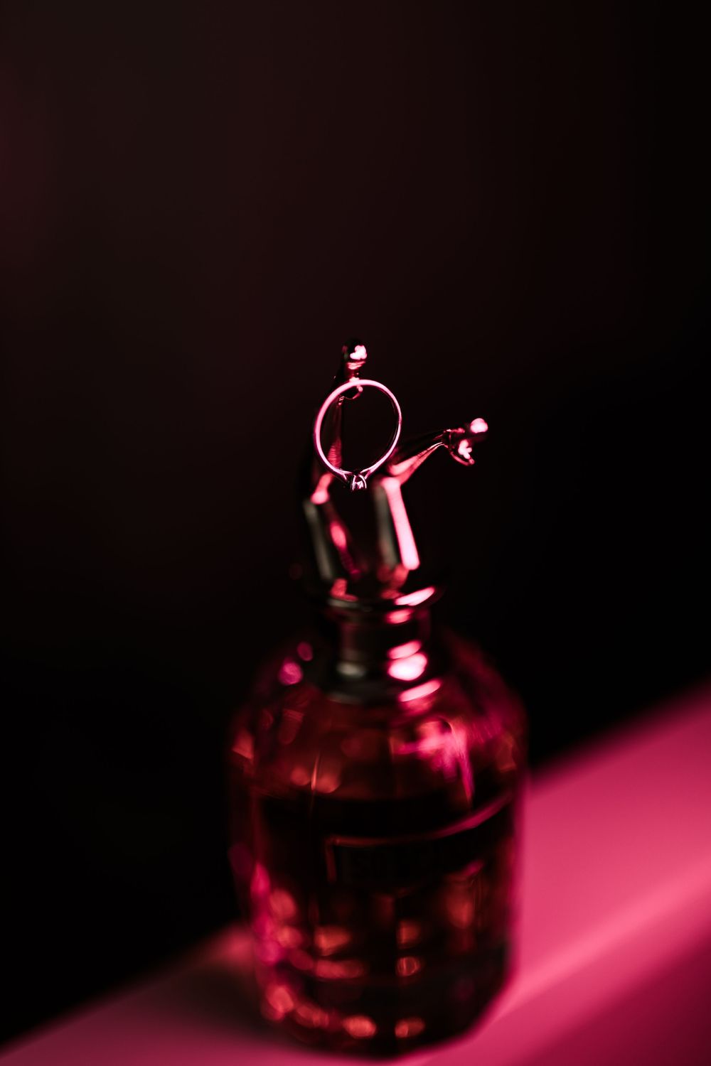 Luxury perfume bottle with wedding rings balanced on dropper in dramatic pink and black lighting.