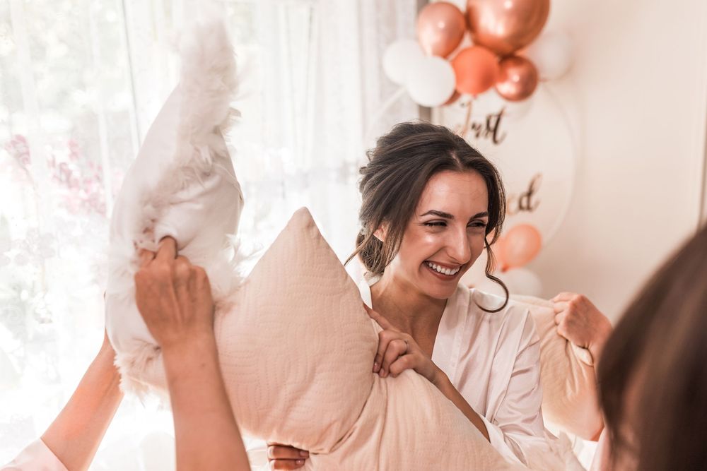 Joyful bride in white robe having a playful pillow fight at a rose gold bridal shower party.