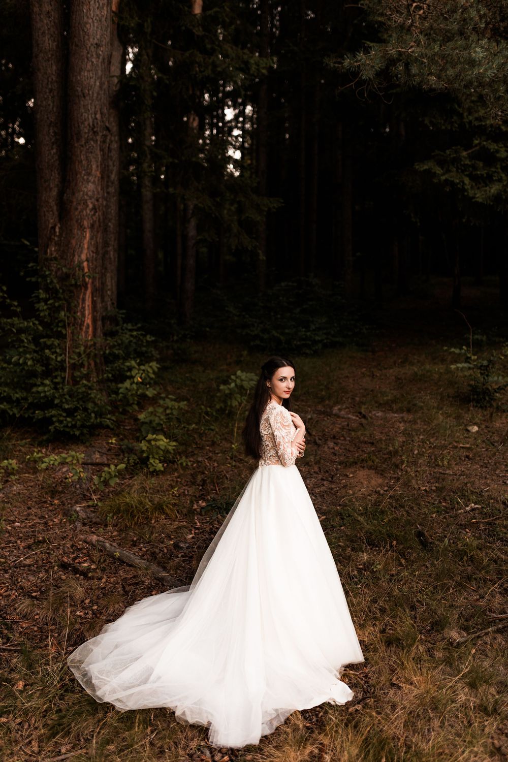 Bride in white lace ballgown with long train standing in dark forest looking over shoulder.
