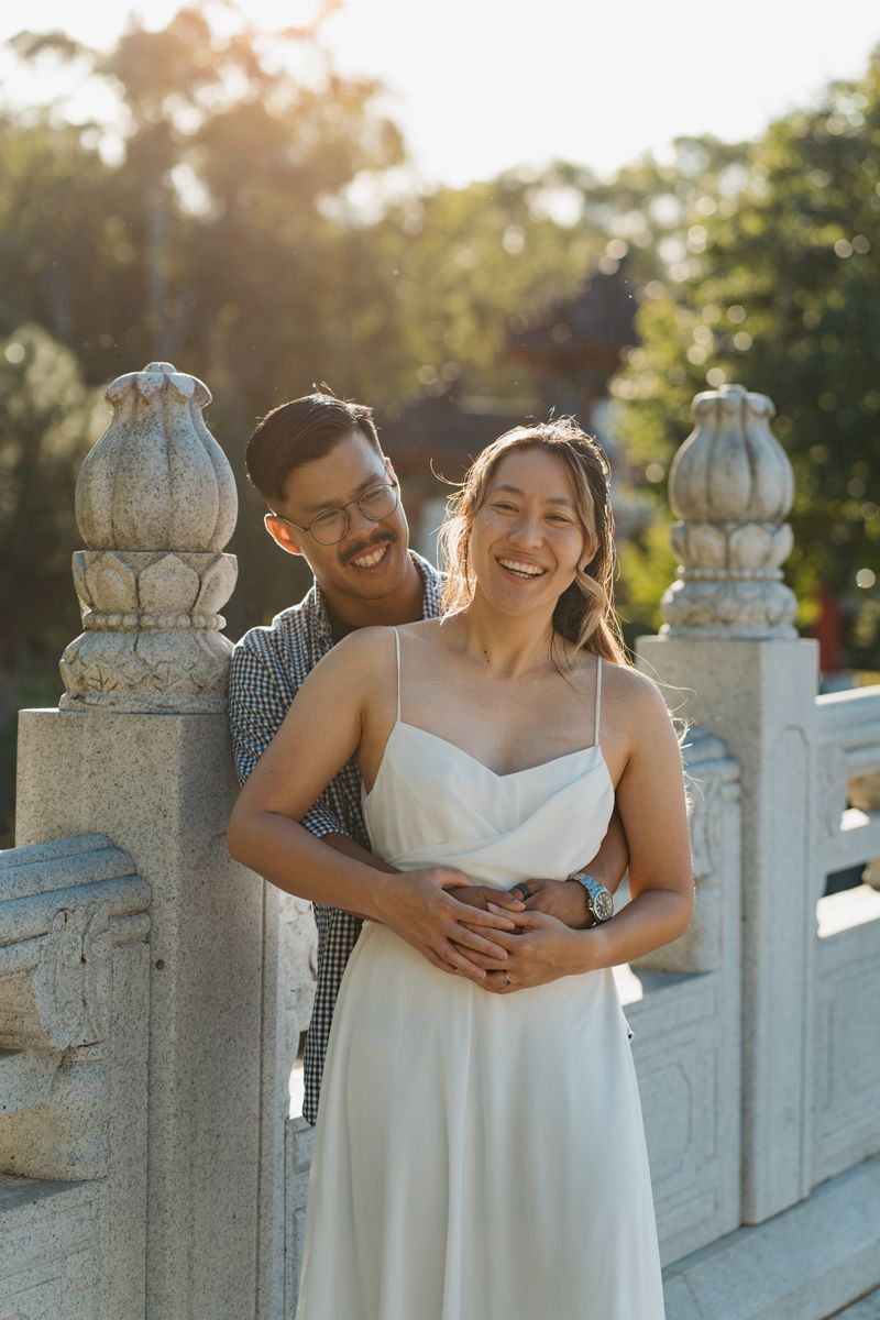 Happy Asian couple embracing on a stone bridge, woman in white dress, man in plaid shirt, golden sunlight backdrop couples photoshoot in syd