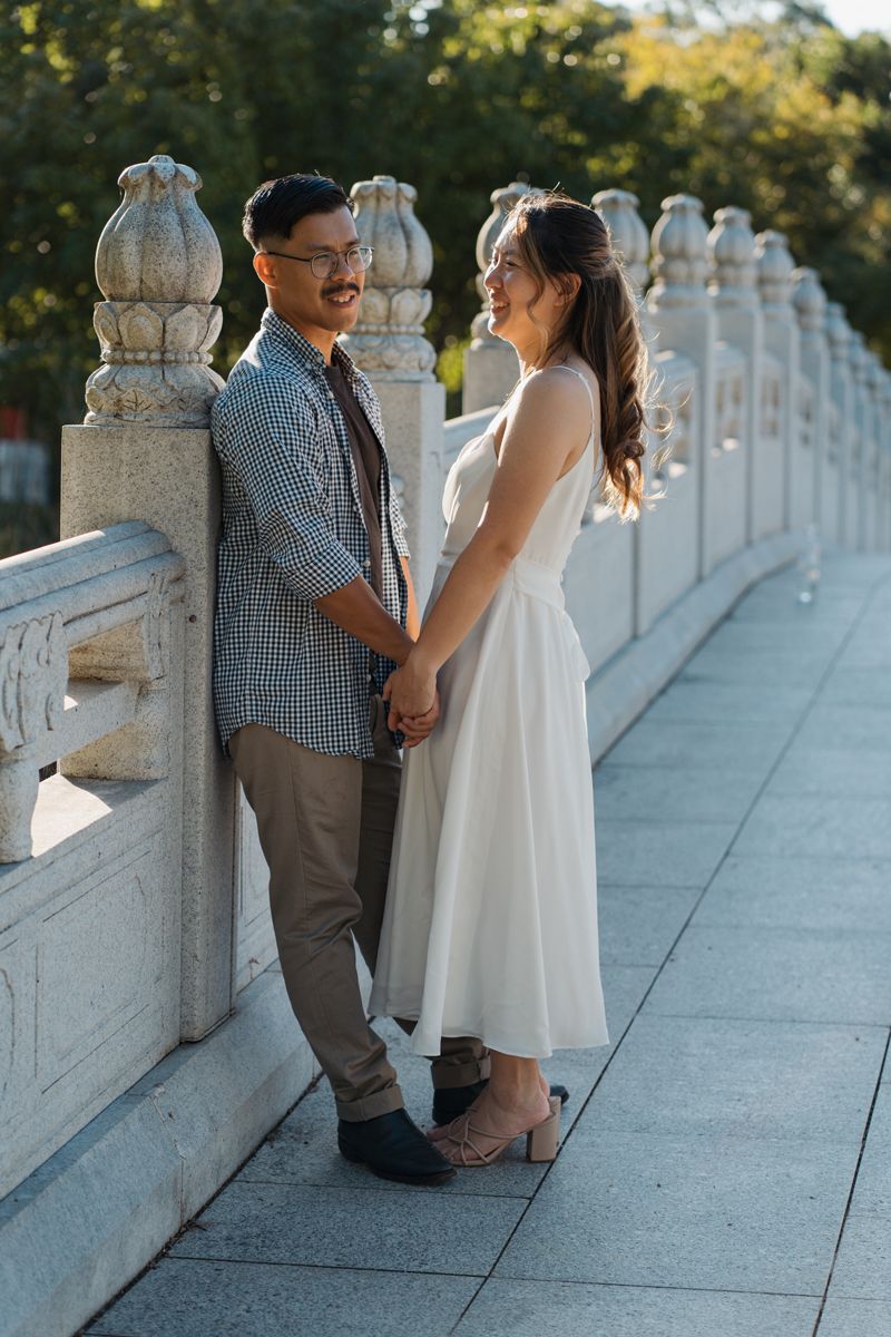 A smiling couple holds hands on a stone bridge walkway, woman in white dress, man in plaid shirt, surrounded by greenery engagement photos
