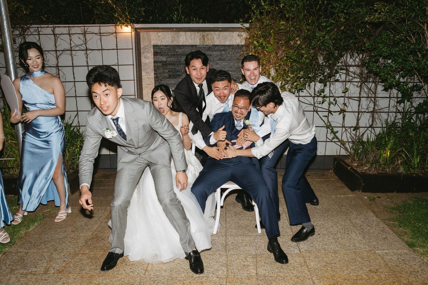 Wedding party playfully lifting groom outdoors at night, bride in white gown, groomsmen in suits laughing.