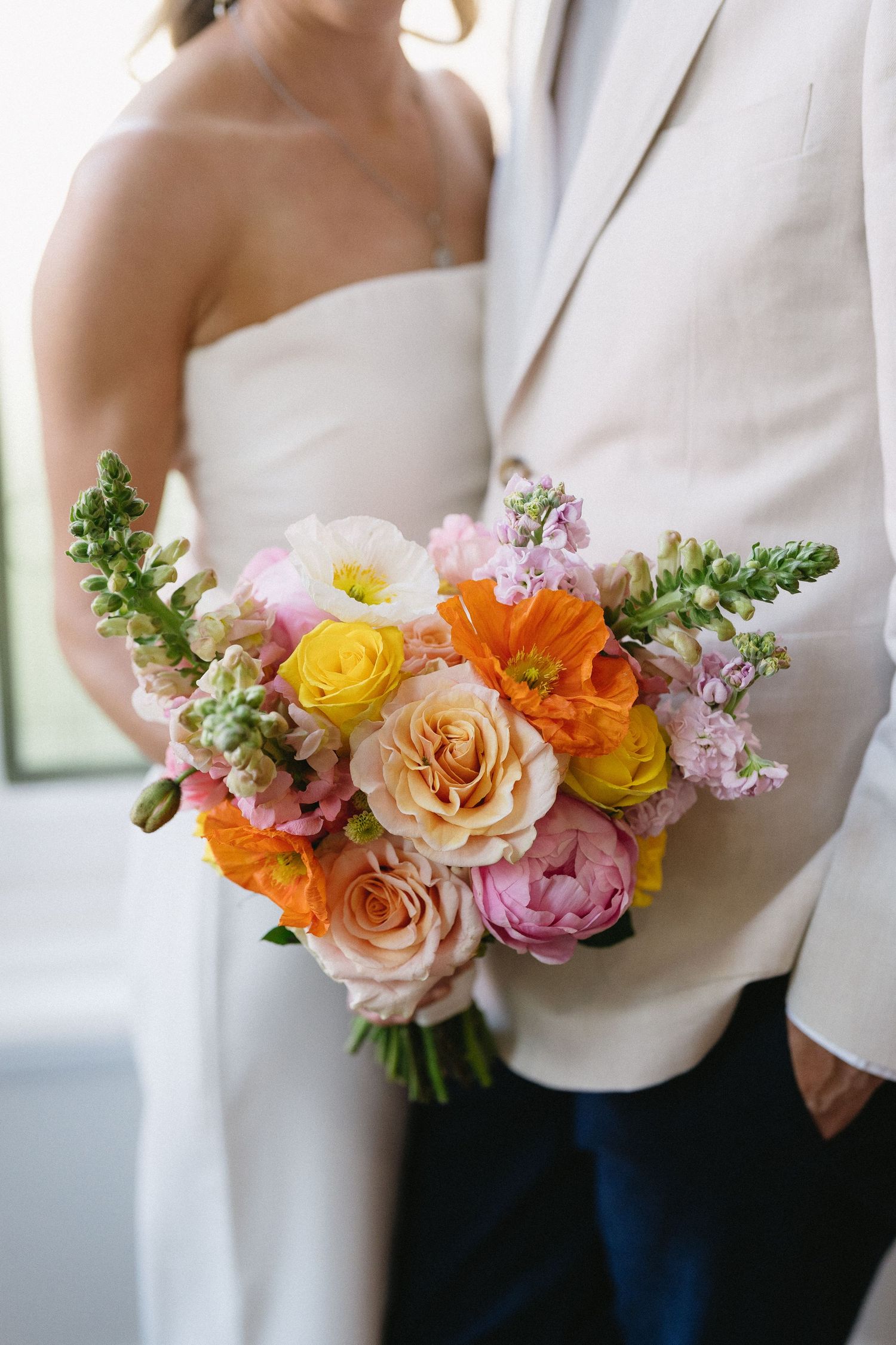 Bride holding a vibrant wedding bouquet with orange, yellow, pink, and peach roses and wildflowers.