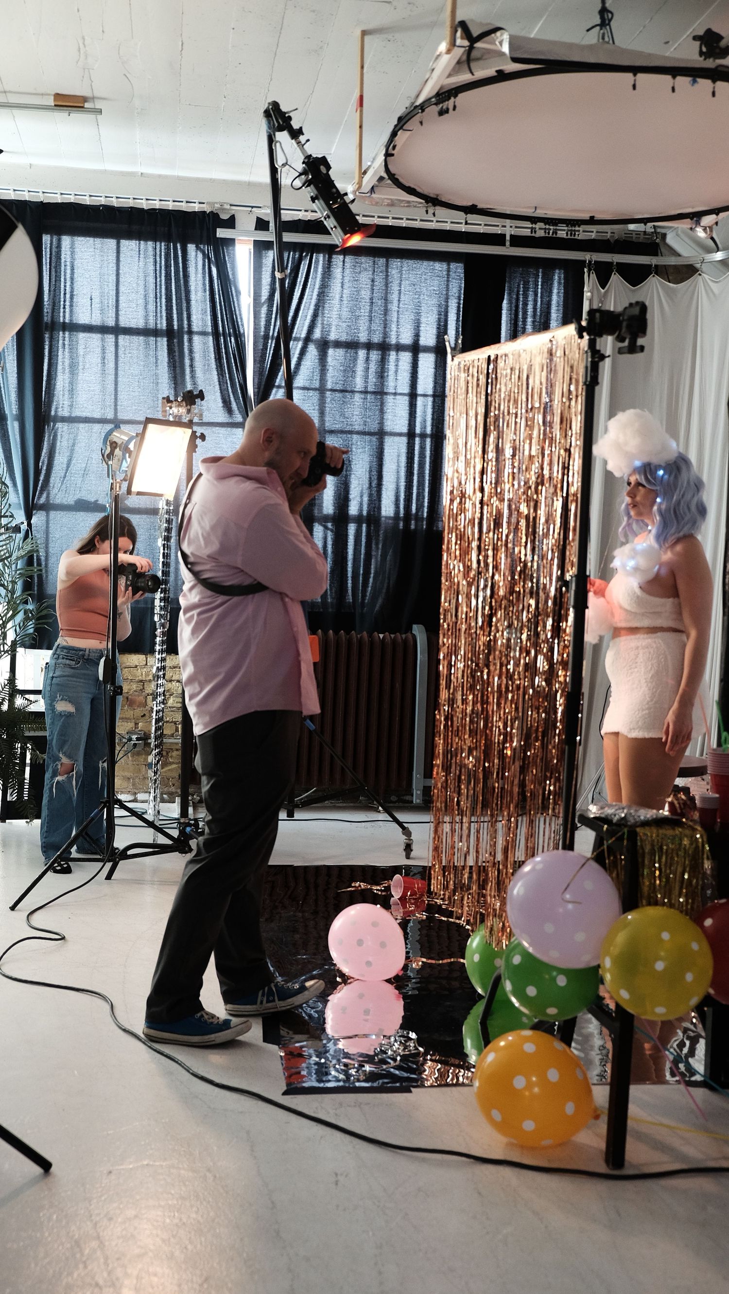 Photographer capturing a model in a studio with balloon decor, tinsel backdrop, and professional lighting equipment.