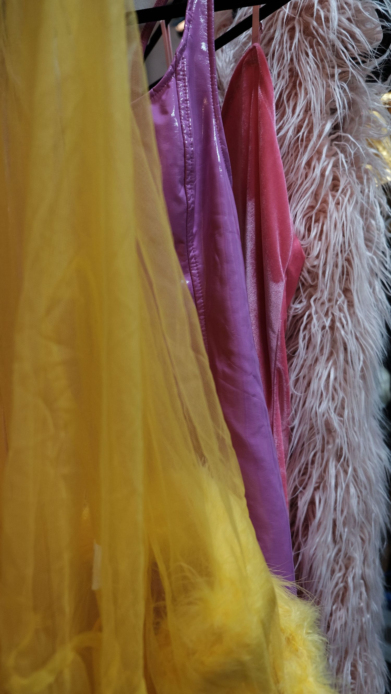 Colorful garments on a rack featuring yellow tulle, purple satin, red dress, and white feather boa in a boutique.
