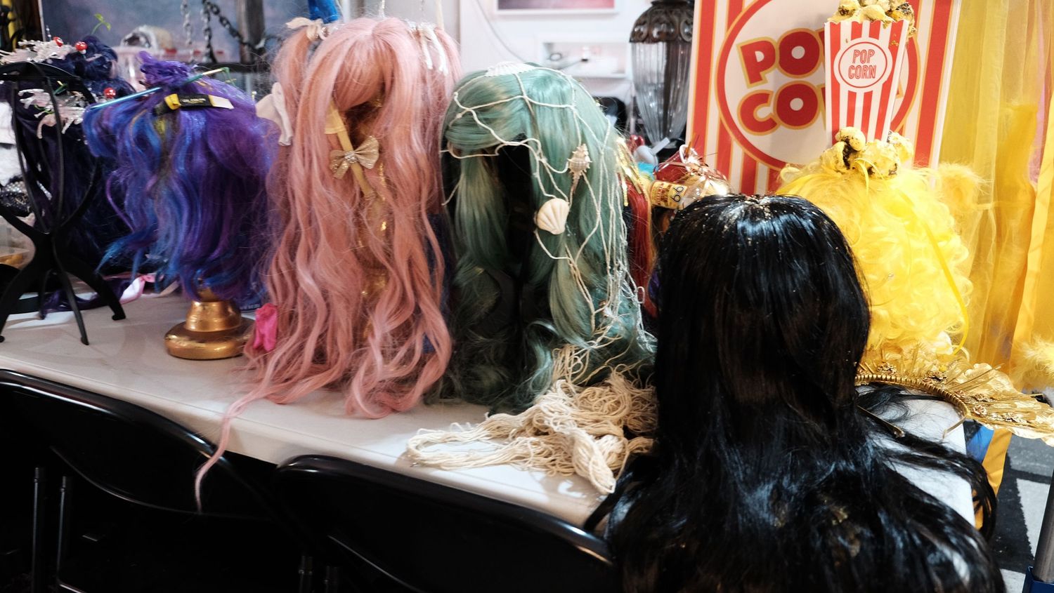 Colorful costume wigs on display stands at a convention, featuring pink, teal, and black styles near a popcorn sign.