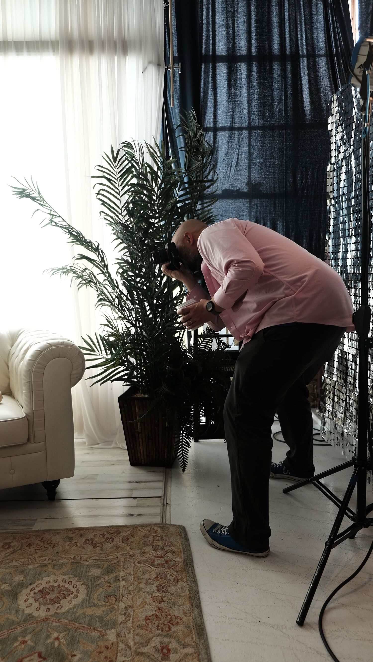 Photographer leaning over a potted palm plant in a bright studio with a white sofa and dark curtain backdrop.