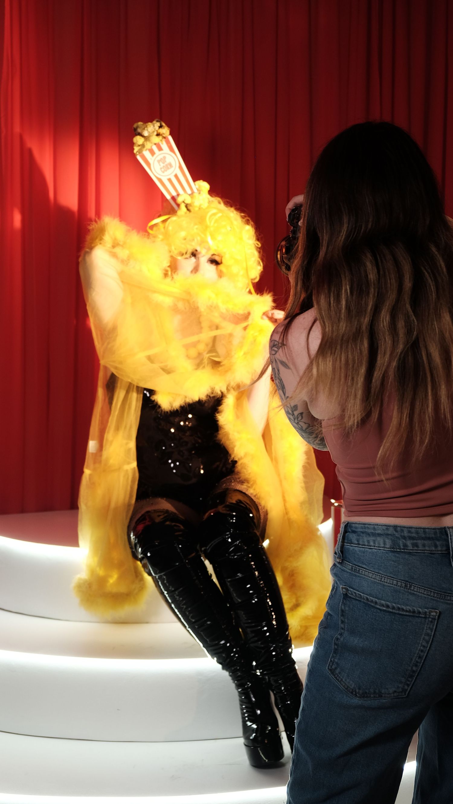 Woman photographing a flamboyant drag queen in yellow feathers and black sequin outfit with popcorn headpiece.