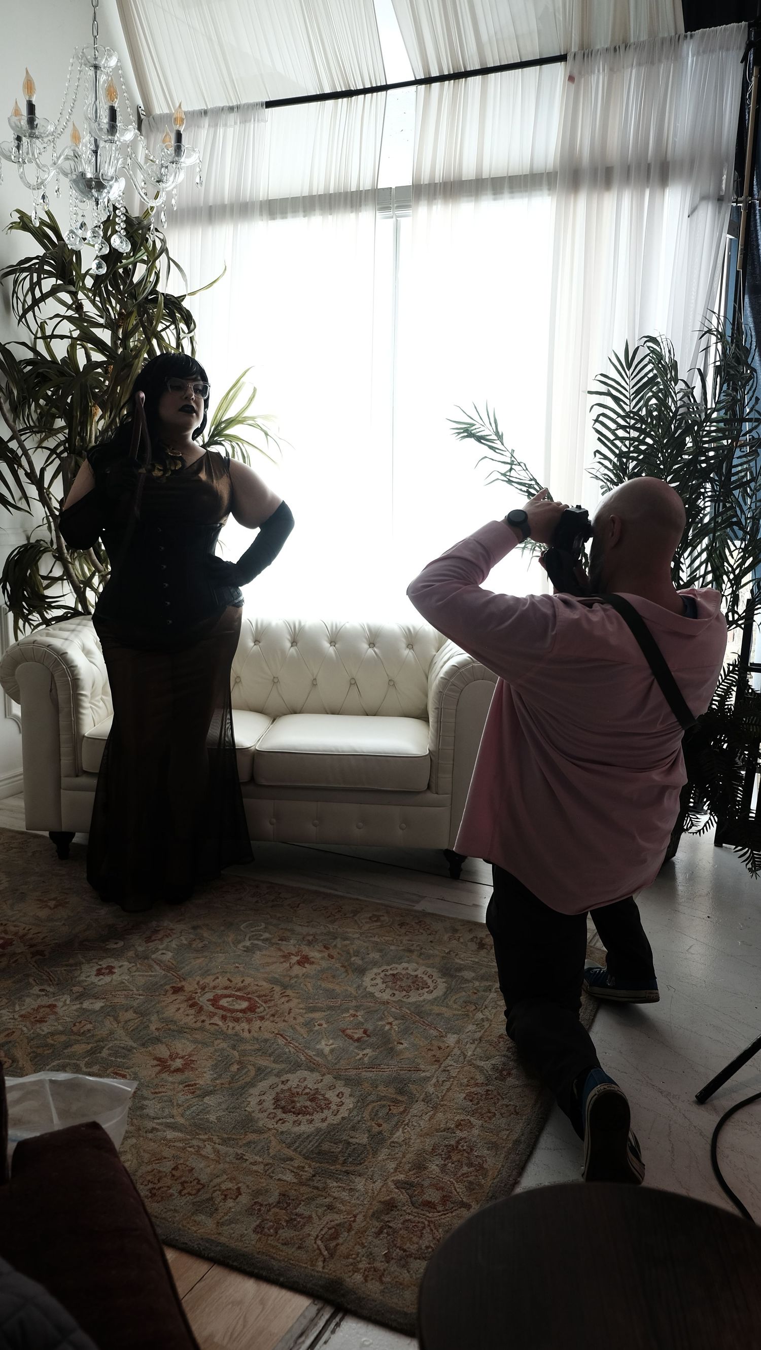 Photographer capturing a woman in elegant black gown posing in a bright studio with white sofa and tropical plants.