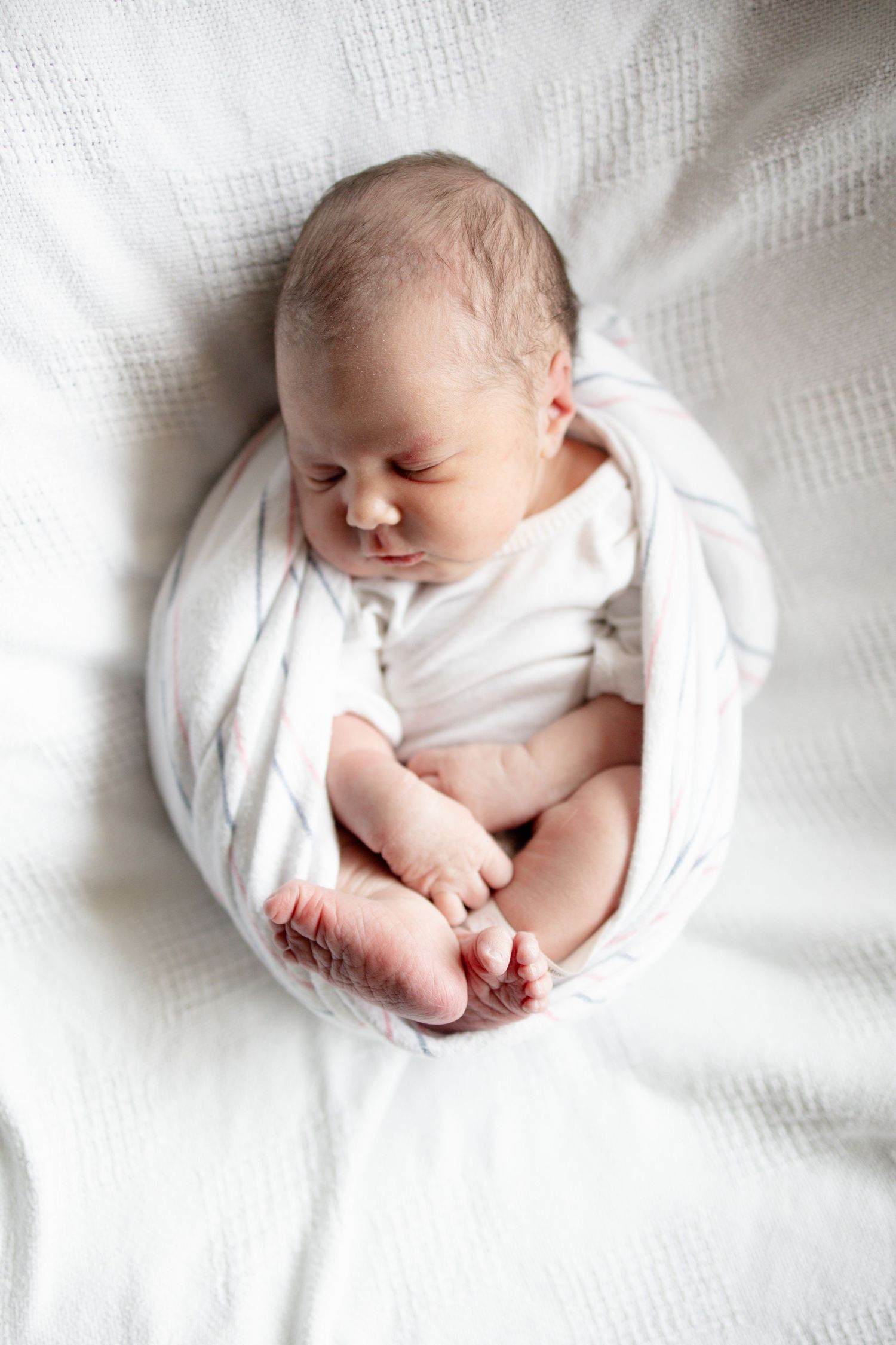 Newborn baby swaddled in white wrap sleeping peacefully on a soft white blanket with tiny feet visible.