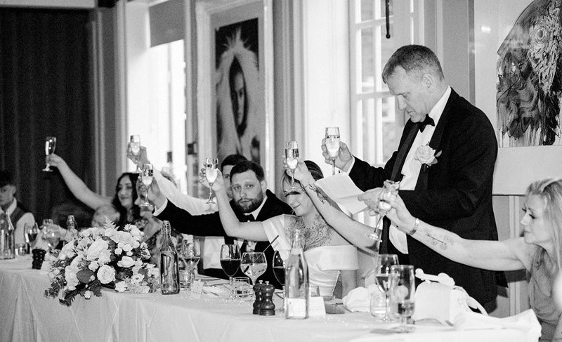 Black and white wedding reception toast with guests raising glasses around a floral centerpiece table.