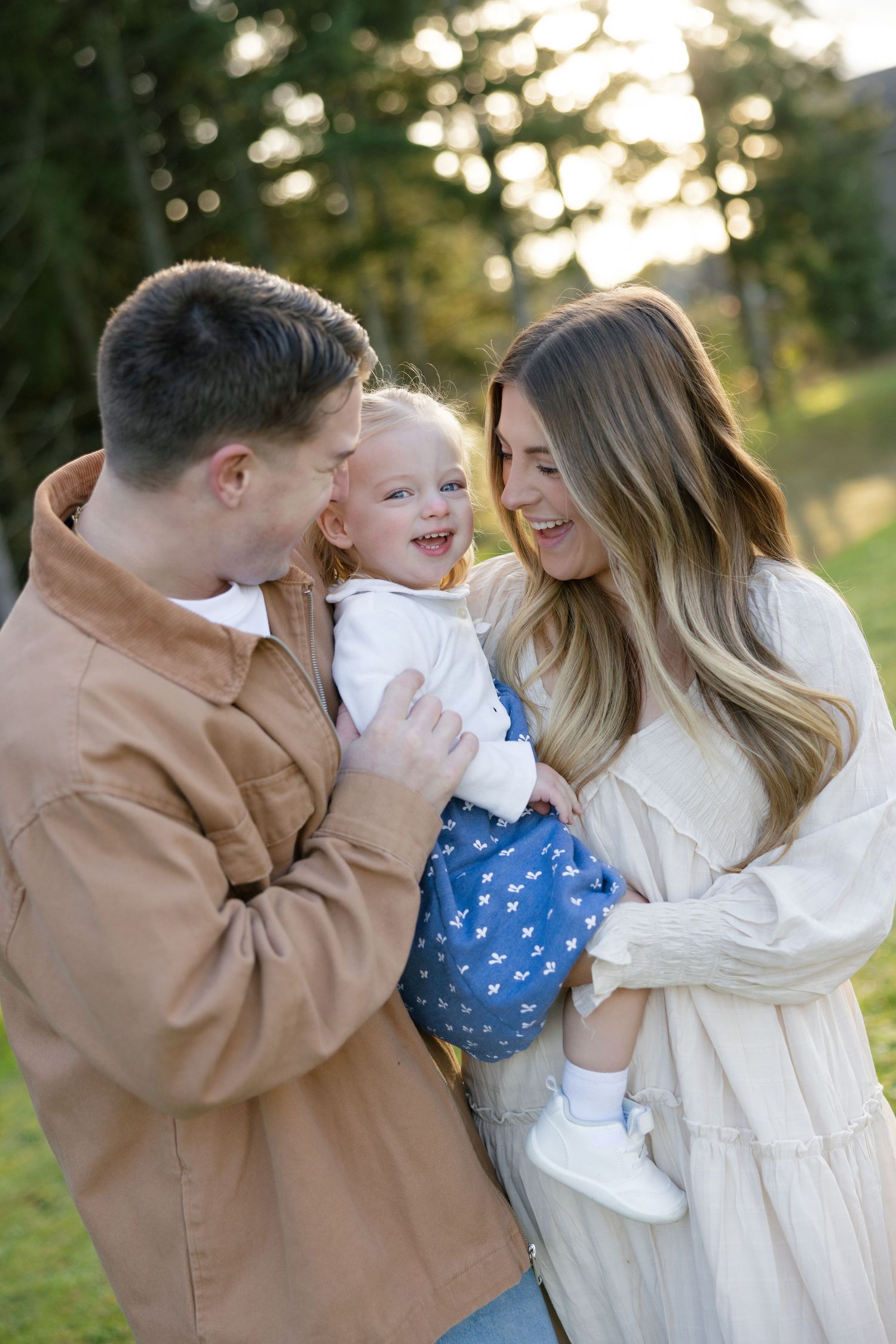 Happy young family with toddler laughing together outdoors in warm golden sunlight.