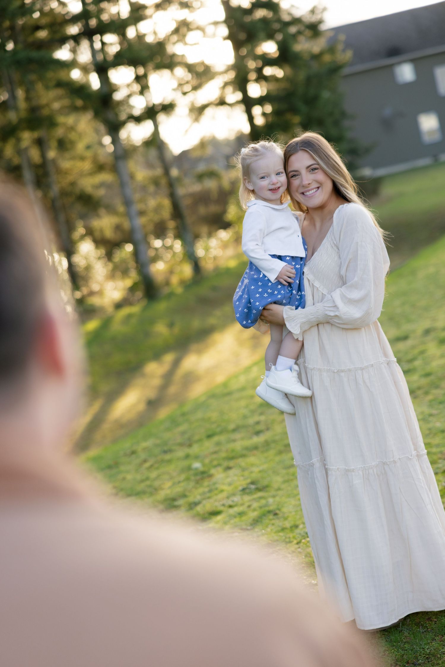 Smiling mother in cream dress holds laughing toddler while father looks on in sunny park.