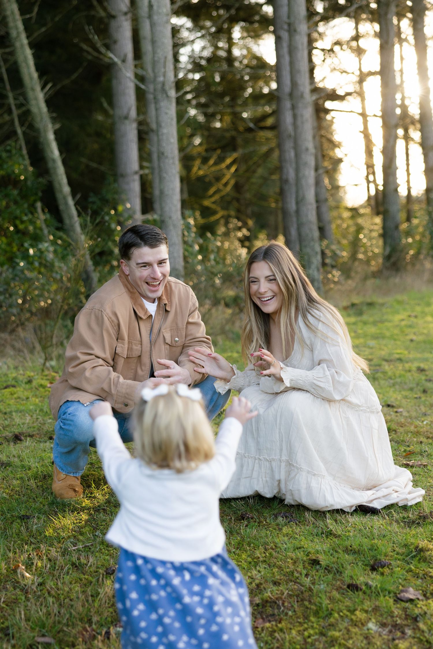 Laughing parents crouch on grass as their toddler daughter walks toward them in forest setting.