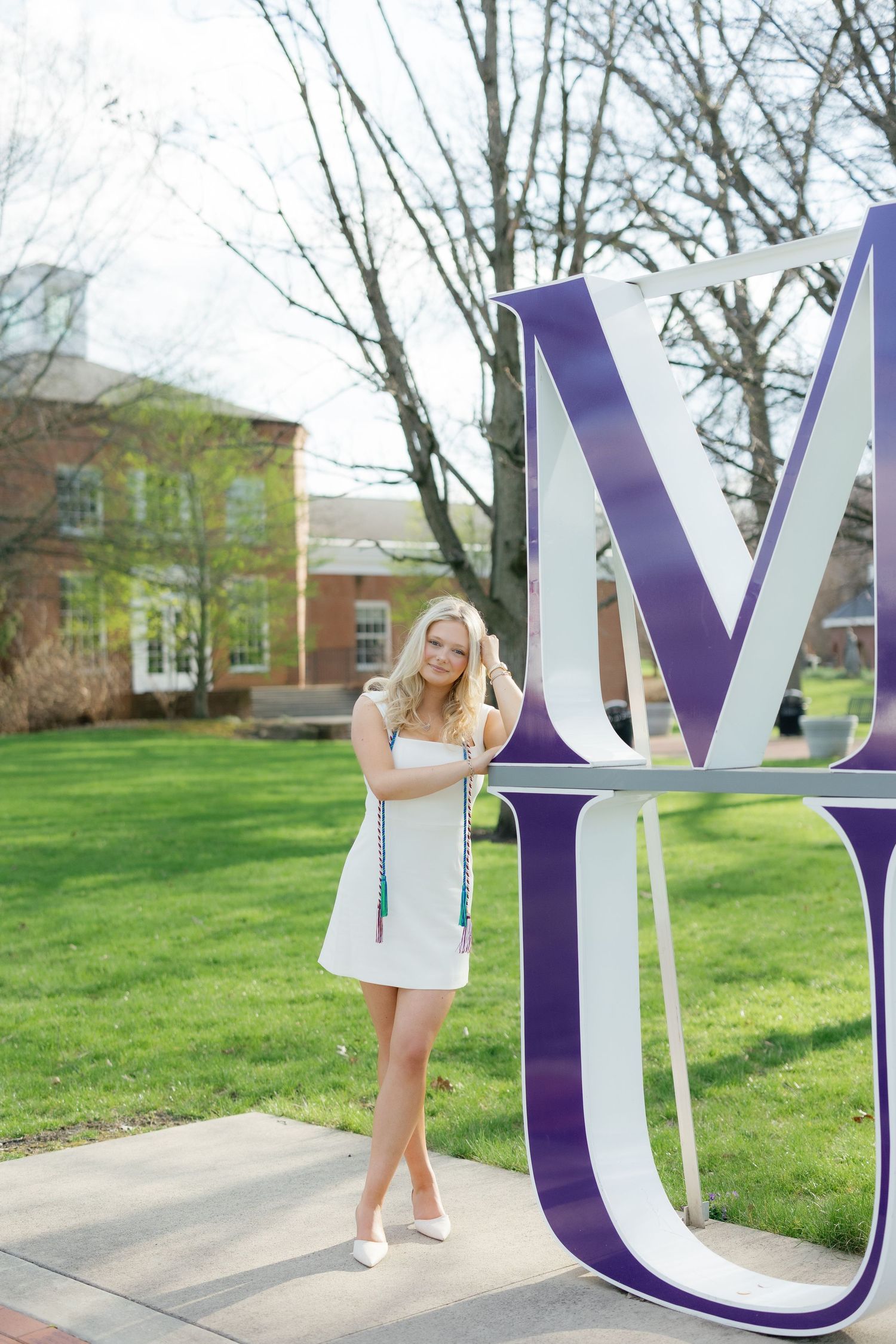 Blonde woman in white dress poses by large purple MU letters on a college campus lawn.