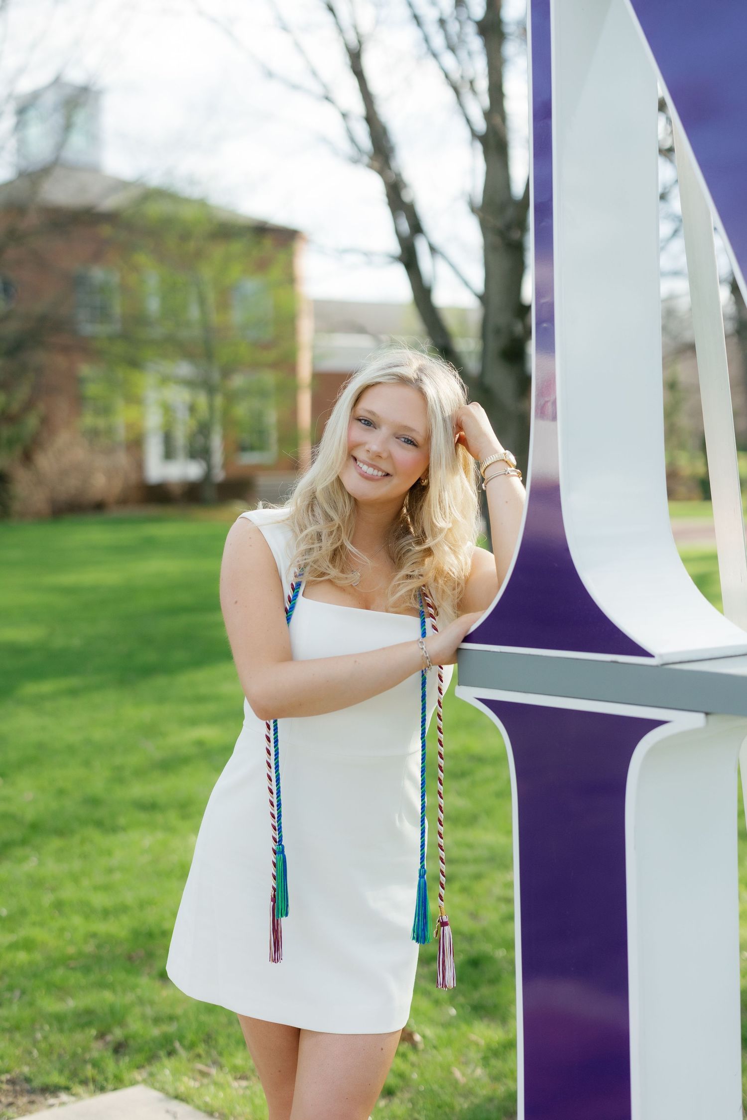 Smiling blonde graduate in white dress with honor cords leans against purple university sign.