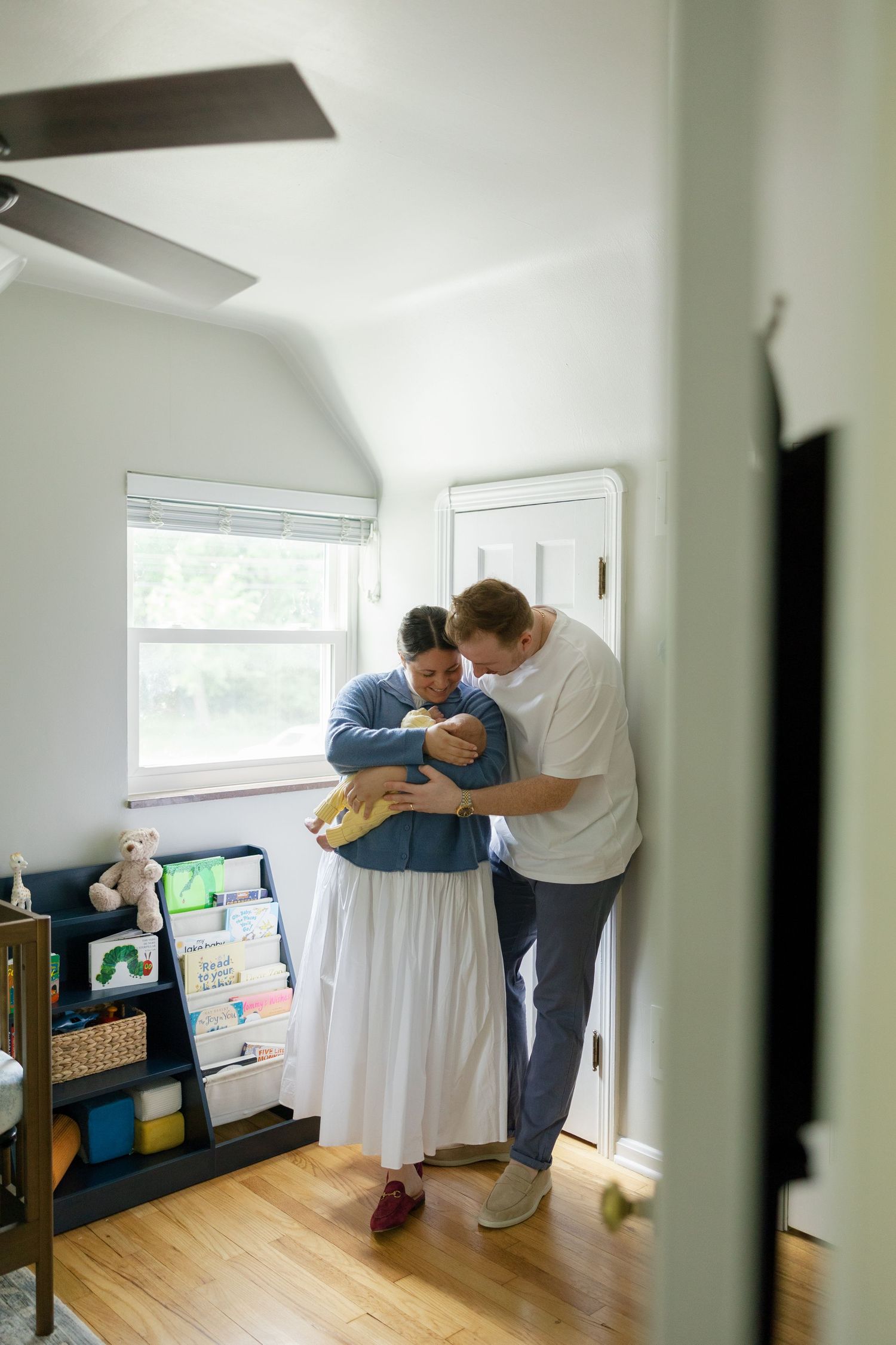 Two parents lovingly cradle their newborn baby together in a softly decorated nursery room with wooden floors.
