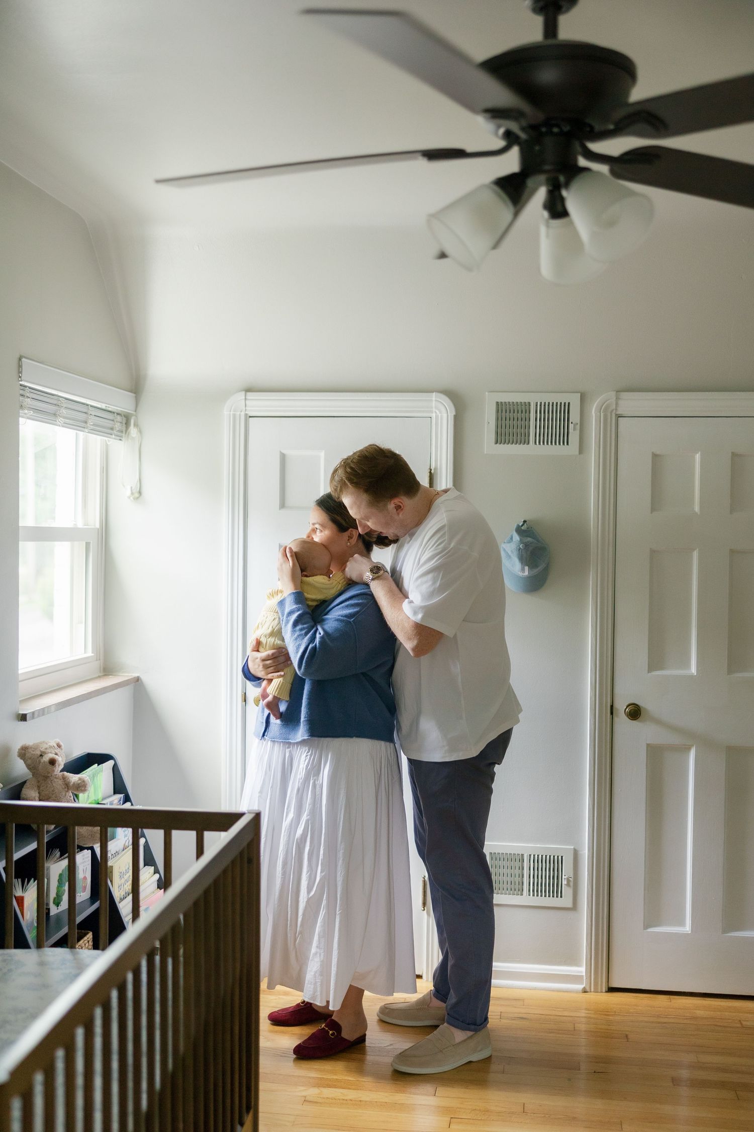New parents share a tender embrace while holding their newborn baby in a warmly decorated nursery with a ceiling fan.