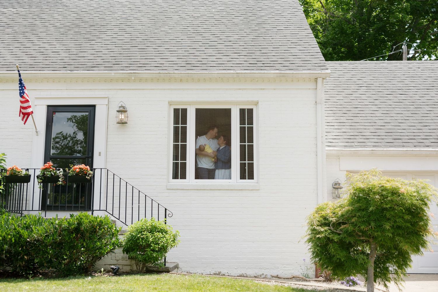 A person holds a newborn baby while standing inside a white house window surrounded by lush green landscaping.