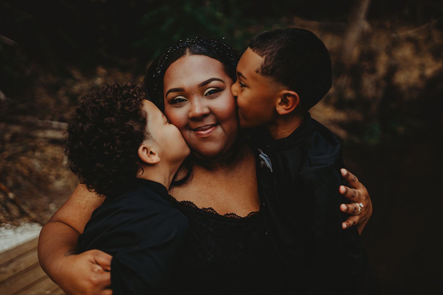 A joyful mother in a black dress receives kisses on both cheeks from her two young sons outdoors.