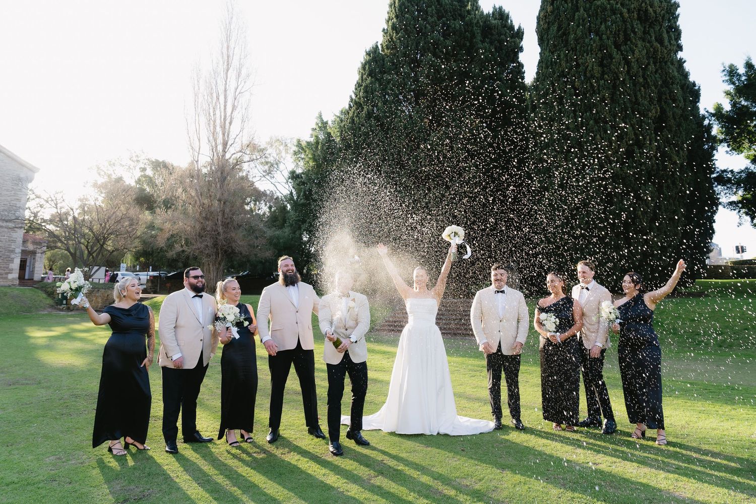 Wedding party poses in elegant courtyard, bride in white gown with groomsmen in black suits and bridesmaids in blush dresses.