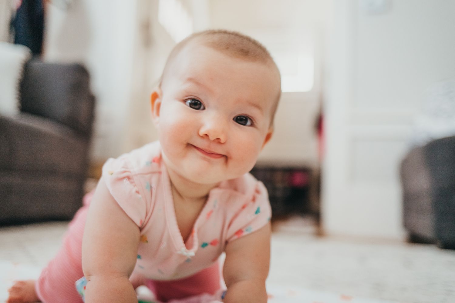 Adorable baby girl in pink onesie smiling while crawling on floor in bright living room.