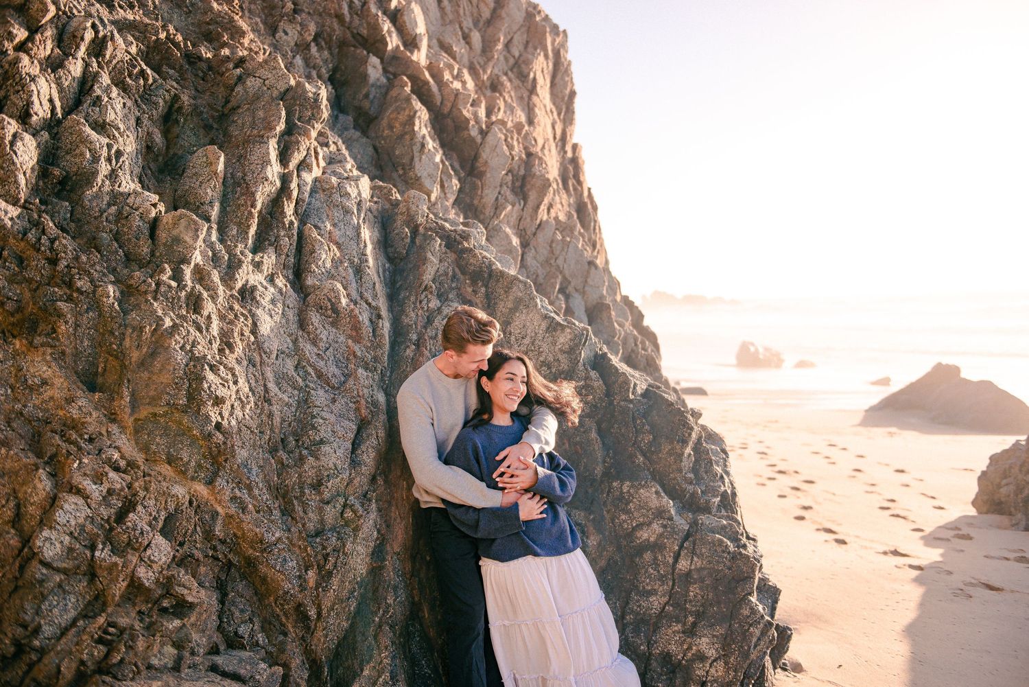 A couple embraces tenderly against a rocky coastal cliff on a sandy beach at golden hour.