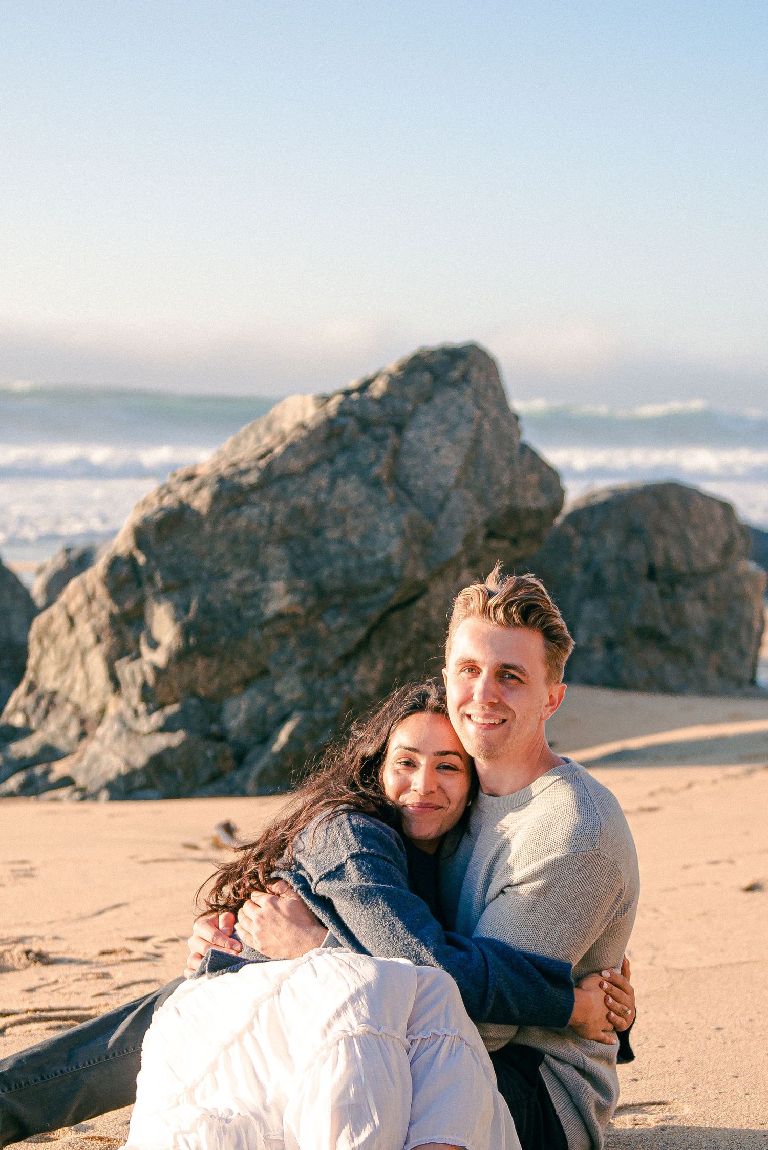 A happy couple embraces on a sandy beach with large rocks and ocean waves in the background.