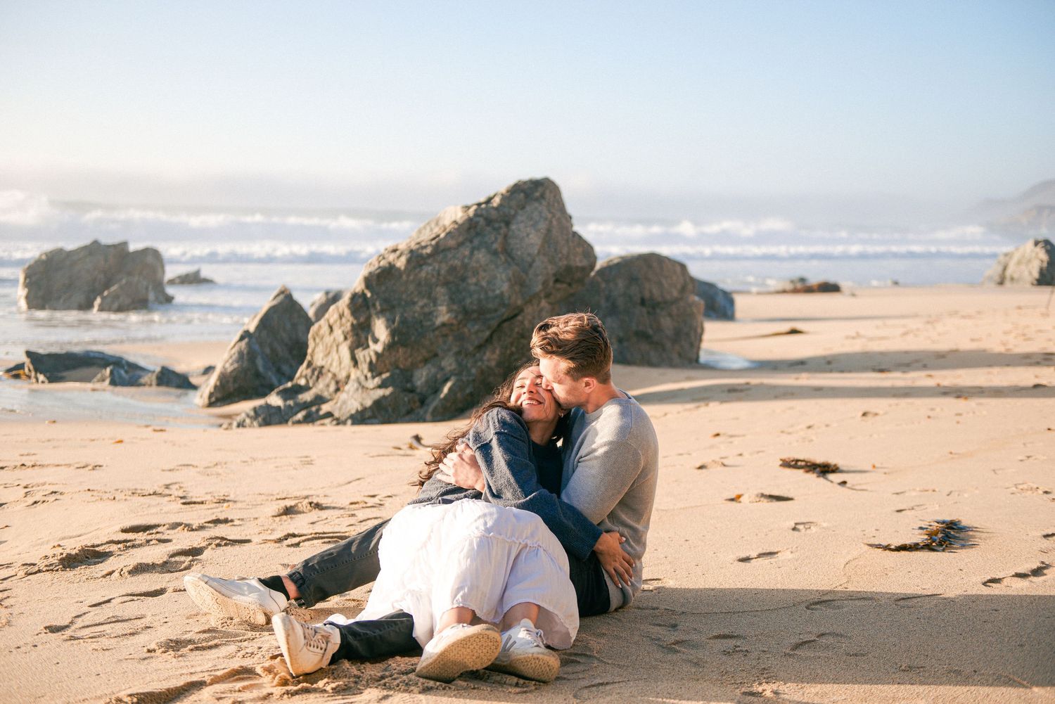 Couple sitting together on sandy beach near large rocks, sharing a romantic embrace by the ocean.